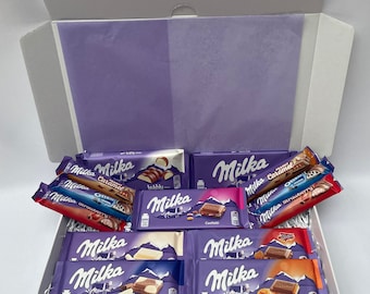 May include: A white gift box filled with various Milka chocolate bars. The bars are in purple packaging with different flavors like caramel, Oreo, and strawberry. The box is lined with purple tissue paper.
