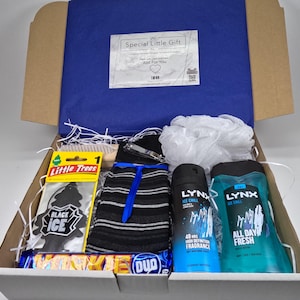 May include: A white gift box filled with various items, including a black and white striped sock, a black car air freshener, a blue and white shower gel bottle, a blue and white body spray bottle, a white bath pouf, a wooden brush, a silver nail clipper, and a chocolate bar.