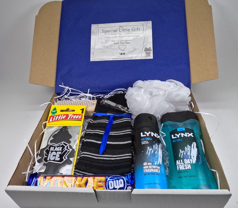May include: A white gift box filled with various items, including a black and white striped sock, a black car air freshener, a blue and white shower gel bottle, a blue and white body spray bottle, a white bath pouf, a wooden brush, a silver nail clipper, and a chocolate bar.