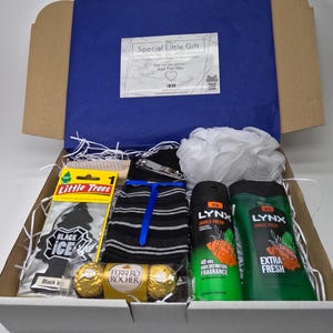 May include: A white gift box filled with various items, including a black and white striped sock, a black ice car air freshener, a black and white shower pouf, two Lynx body wash bottles, two chocolate candies, and a small wooden comb.