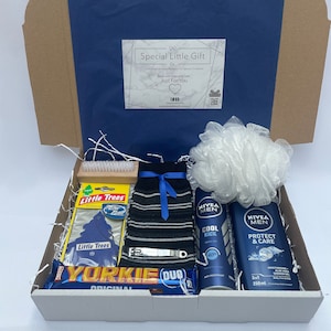 May include: A gift box filled with various items, including a blue and white striped sock, a white bath puff, a bottle of Nivea Men Cool Kick deodorant, a bottle of Nivea Men Protect & Care body wash, a small wooden brush, a car air freshener, and a chocolate bar. The box is lined with white shredded paper.