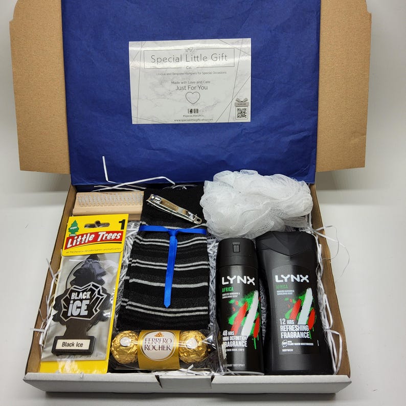 May include: A gift box filled with various items, including a pair of black and white striped socks, a white bath pouf, two black bottles of Lynx Africa body spray, a car air freshener, two chocolate candies, a nail clipper, and a wooden brush. The box is lined with white shredded paper.