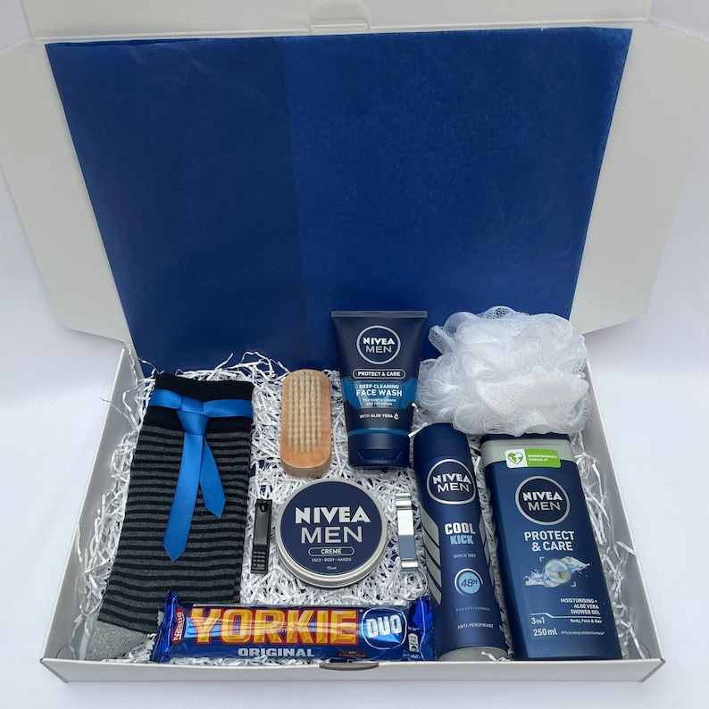May include: A blue and white gift box filled with men's grooming products, including face wash, body wash, shaving cream, and a loofah. The box also includes a pair of striped socks, a wooden brush, a chocolate bar, and a metal razor.