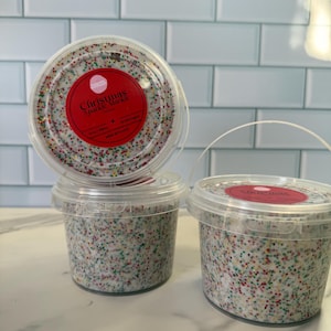May include: Three clear plastic containers filled with colorful sprinkles. The containers have red labels that read "Christmas Sparkle Mix". The sprinkles are a mix of red, green, yellow, and white.