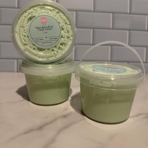 Puede incluir: Tres recipientes de plástico transparente con slime verde, uno apilado encima de otro. El recipiente superior tiene una tapa con una etiqueta que dice "Marshmallow Fluff Salad" y "Slime". Los recipientes están sobre una superficie de mármol blanco.