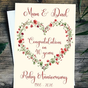 May include: A cream-colored greeting card featuring a heart-shaped wreath of red flowers and green foliage. The text reads: Mum & Dad, Congratulations on 40 years, Ruby Anniversary, 1986-2026. The card is displayed against a dark wooden background.