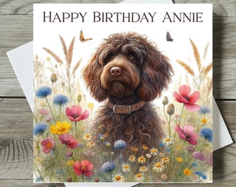 May include: A greeting card with the text "HAPPY BIRTHDAY ANNIE" features a brown dog in a field of colorful flowers. The dog wears a brown collar. Butterflies are also present in the image.