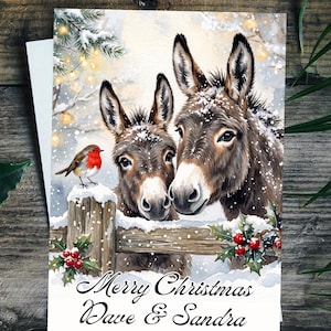 Könnte beinhalten: Eine Weihnachtsgrußkarte mit einer Aquarellillustration von zwei braunen Eseln und einem Rotkehlchen auf einem verschneiten Holzzaun mit Stechpalmenbeeren. Der Text lautet Merry Christmas Dave & Sandra in schwarzer Schreibschrift.