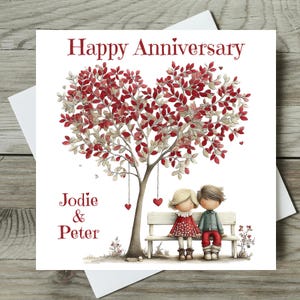 May include: A white anniversary card with the words "Happy Anniversary" in red. The card features a tree with red and white leaves shaped like a heart, with two figures sitting on a bench. The names "Jodie & Peter" are also on the card.