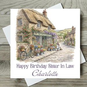 Könnte beinhalten: Eine Aquarell-Geburtstagskarte mit einem Blumengeschäft, "The Wild Daisy", mit Strohdach und Blumenschmuck. Ein Fahrrad mit Korb steht davor. Die Karte trägt die Aufschrift "Happy Birthday Sister In Law Charlotte."