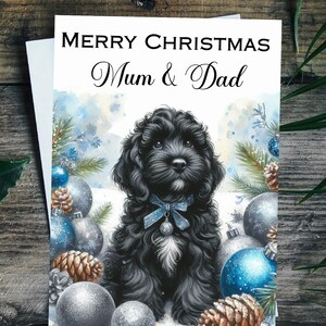 May include: Christmas card with a black dog wearing a blue bow, set amongst baubles and pine cones. The card reads "MERRY CHRISTMAS Mum & Dad" in script. The card is on a wooden surface, with a snowy background.