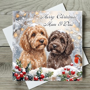 May include: A square Christmas greeting card featuring a watercolour illustration of a golden-brown dog and a dark brown dog sitting in snow with holly berries and a robin. The text reads Merry Christmas Mum & Dad in black cursive script.