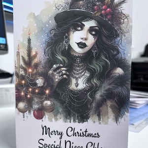 K&ouml;nnte beinhalten: Weihnachtskarte mit einer gotischen Illustration einer Frau mit Zylinder, langen dunklen Haaren und aufwendigem Schmuck. Links ein kleiner Weihnachtsbaum mit Kugeln und Kerzen. Der Text lautet "Merry Christmas Special Niece Chloe."