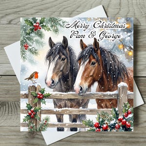 Könnte beinhalten: Eine quadratische Weihnachtsgrußkarte mit einer Aquarellillustration von zwei Pferden hinter einem Holzzaun. Der Text lautet Merry Christmas Pam & George. Ein kleines Rotkehlchen sitzt auf dem Zaun, umgeben von Stechpalmen, Kiefernzweigen und Schnee.