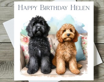 May include: A greeting card with the text "Happy Birthday Helen" features watercolor illustrations of two dogs, one black and one golden brown, seated on a floral patterned couch with blue pillows. The card is on a white background.
