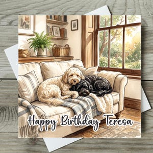 May include: A greeting card featuring a watercolour illustration of a cream-coloured dog and a black dog sleeping together on a beige sofa with a blue and white striped throw blanket. The text Happy Birthday Teresa is written in black script.
