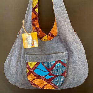 May include: A denim shoulder bag with a colorful patterned fabric pocket and strap accents. The bag features a rectangular pocket with a vibrant design of blue, yellow, and orange shapes. The strap and top edge are lined with the same patterned fabric.