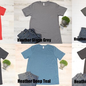 May include: Six different colored t-shirts laid flat on a light wood surface. The colors are heather red, heather storm grey, heather military green, heather midnight navy, heather deep teal, and heather dark grey.