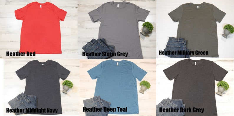 May include: Six different colored t-shirts laid flat on a light wood surface. The colors are heather red, heather storm grey, heather military green, heather midnight navy, heather deep teal, and heather dark grey.