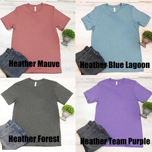 May include: Four plain t-shirts in different colors: heather mauve, heather blue lagoon, heather forest, and heather team purple. Each shirt is laid flat on a white surface with a pair of jeans and a small potted plant.