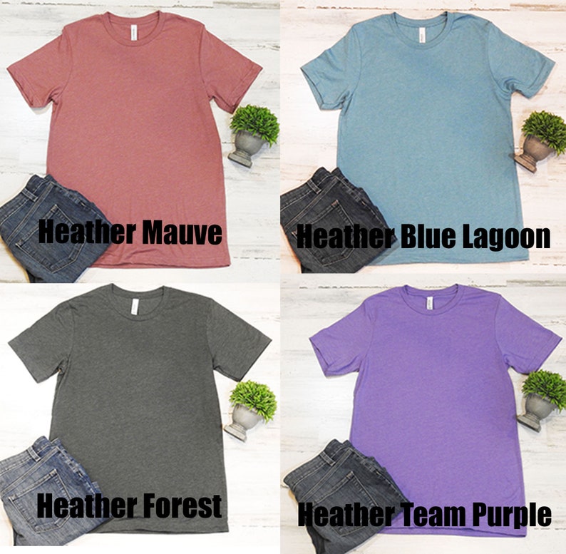 May include: Four plain t-shirts in different colors: heather mauve, heather blue lagoon, heather forest, and heather team purple. Each shirt is laid flat on a white surface with a pair of jeans and a small potted plant.