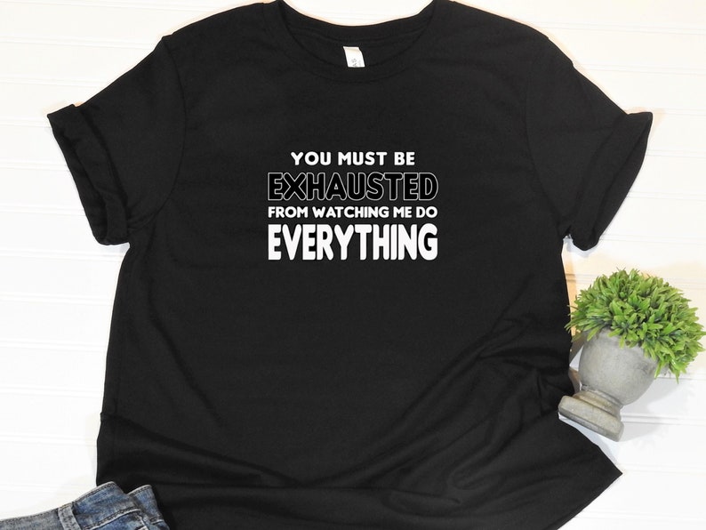 May include: Black t-shirt with white text that reads "You must be exhausted from watching me do everything."