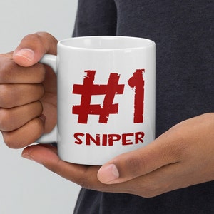 Może przedstawiać: Biały ceramiczny kubek do kawy z czerwonym nadrukiem #1 SNIPER. Kubek jest trzymany przez rękę.