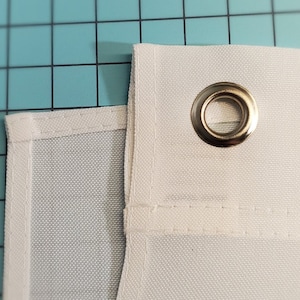 May include: Close-up of white fabric with a metal grommet. The fabric has a textured weave and is stitched along the edges. The grommet is silver and reinforces a hole in the fabric, likely for hanging.