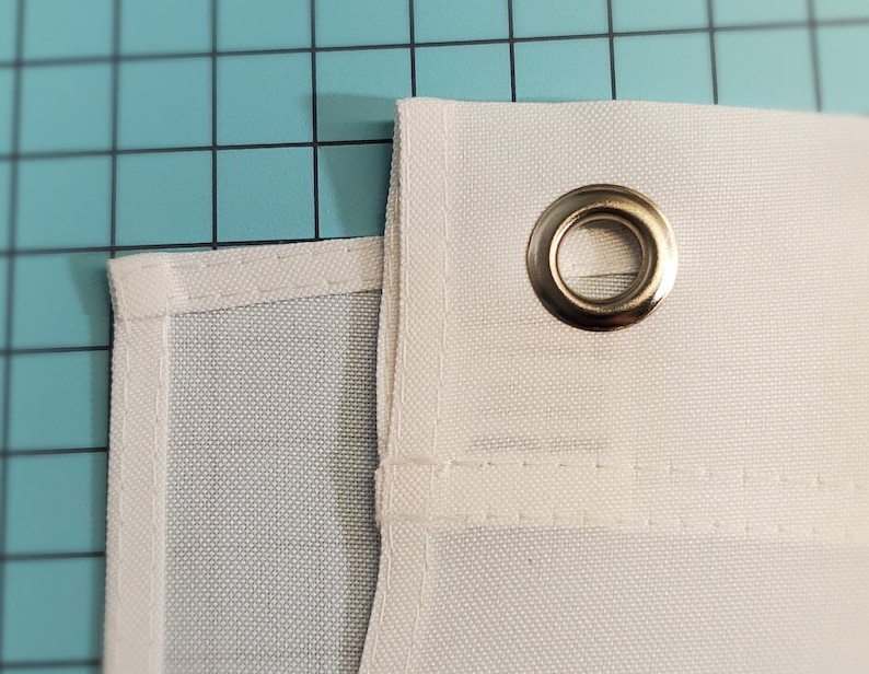 May include: Close-up of white fabric with a metal grommet. The fabric has a textured weave and is stitched along the edges. The grommet is silver and reinforces a hole in the fabric, likely for hanging.