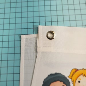 May include: Close-up of a white fabric banner with a cartoon illustration. The banner has a reinforced edge with a metal grommet. The banner is on a blue grid surface.