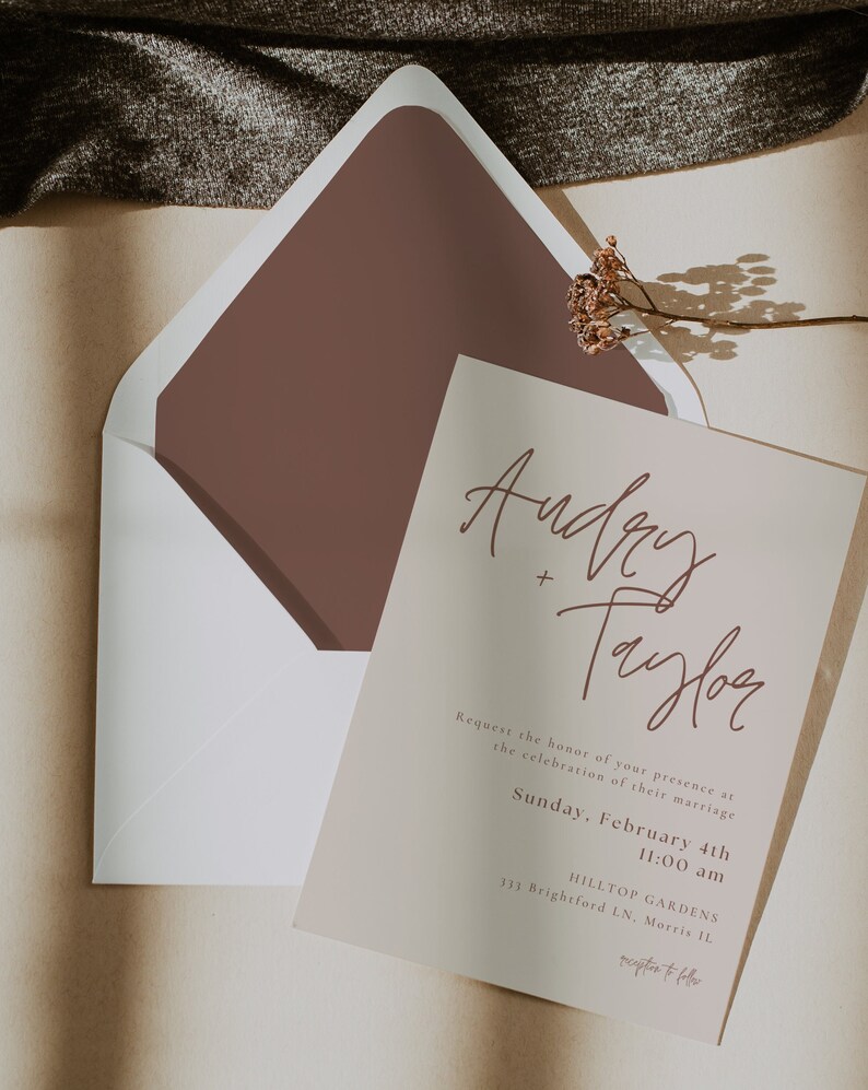 May include: A wedding invitation with a light brown background and a brown envelope. The invitation features the names "Audrey + Taylor" in a handwritten script font. The text reads "Request the honor of your presence at the celebration of their marriage. Sunday, February 4th 11:00 am HILLTOP GARDENS 333 Brightford LN, Morris IL reception to follow"