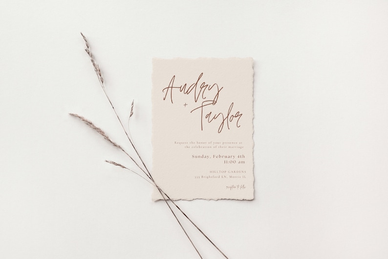 May include: A wedding invitation with a rustic, cream-colored paper texture. The names "Audrey + Taylor" are written in a cursive script in brown ink. The invitation details the date and time of the wedding ceremony, which is Sunday, February 4th at 11:00 am at Hilltop Gardens.