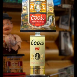May include: A novelty lamp crafted from a Coors beer can, with a lampshade displaying the Coors logo and a mountain landscape. The can base is labelled "America's Fine Light Beer" and "12 Fluid Ounces". A decorative item.