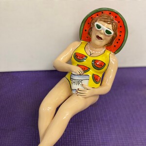 May include: A ceramic figurine of a woman wearing a yellow swimsuit with watermelon print and a watermelon hat. She is holding a container of chips and wearing sunglasses.