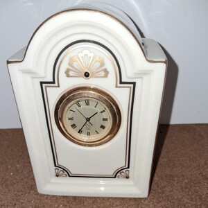 May include: A white porcelain clock with gold accents and a black clock face. The clock is in the shape of a small house with an arched top.