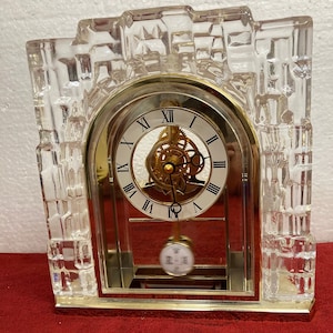 May include: A clear crystal clock with a gold frame and Roman numeral clock face. The clock has a pendulum and is set against a white background.