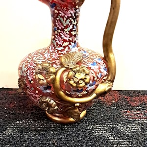May include: A red glass pitcher with a gold handle and floral design. The pitcher has a bulbous body and a flared rim. The floral design includes blue, white, and pink flowers. The handle and decorative accents are gold.