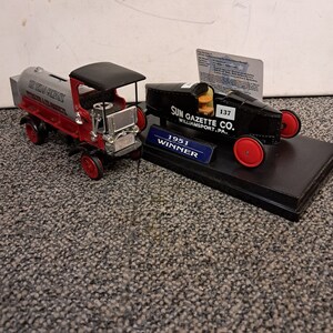 May include: A black and red toy model of a 1951 race car with the text "SUN GAZETTE CO. WILLIAMSPORT, PA. 1951 WINNER" on the side. The car is on a black display base with a smaller toy model of a gray and red tanker truck.