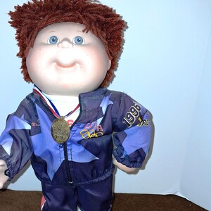 May include: A Cabbage Patch Kid doll wearing a blue and white tracksuit with a gold medal around its neck. The doll has red hair and blue eyes. The tracksuit has the words "USA" and "1996" on it.