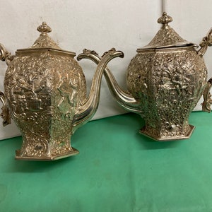 May include: Two silver teapots with ornate designs and handles. The teapots are on a green surface.