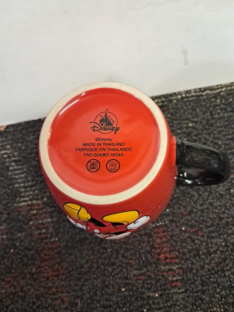 May include: Red ceramic mug featuring Mickey Mouse. The mug has a black handle and the text "&copy;Disney MADE IN THAILAND FABRIQUE EN THAILANDE FAC-024367-18346" printed on the bottom. The mug has a glossy finish.