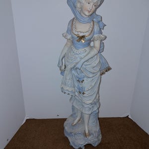 May include: A large porcelain figurine of a woman in a blue dress and hat. She is holding a small object in her hand and is standing on a base.