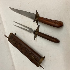 May include: A wooden handled carving knife and fork set with a matching wooden sheath. The handles are intricately carved with floral designs.