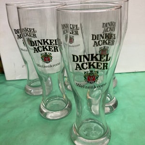 May include: Six clear glass beer mugs with a green tint. Each mug has the text "Dinkel Acker" and a logo with a crest and wheat stalks. The text "Weizenkrone" is printed below the logo.