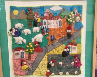 May include: A framed felt artwork depicting a colorful scene with houses, trees, sheep, and people. The artwork is stitched with various colors of thread and includes the text "Casa de la Tia".