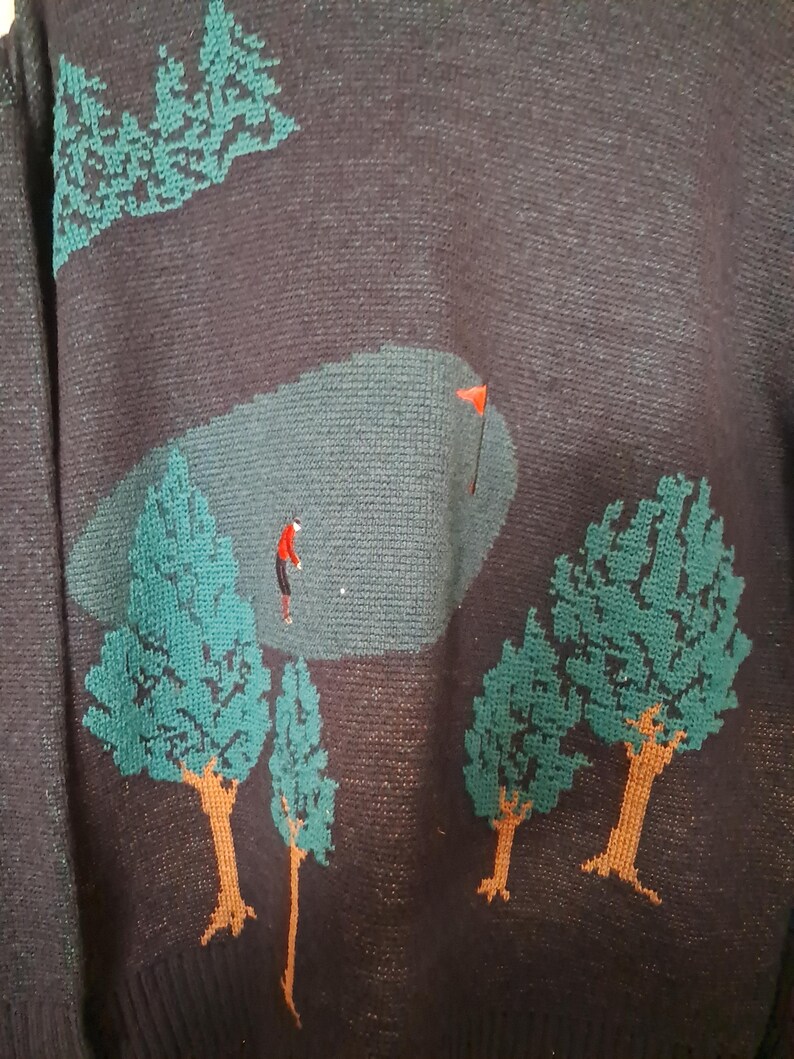 May include: Dark navy blue sweater with a golf course design. The sweater features a green golf course scene with a golfer in red, green trees, and a red flag. The trees have brown trunks and green foliage.
