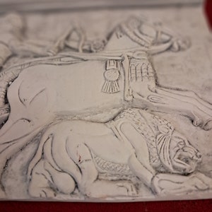 May include: White plaster relief sculpture of a rider on horseback, with a lion lying beneath the horse. The horse is wearing a saddle and bridle with a decorative buckle. The sculpture is in a realistic style.