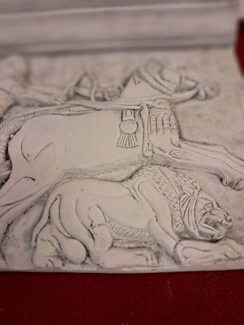 May include: White plaster relief sculpture of a rider on horseback, with a lion lying beneath the horse. The horse is wearing a saddle and bridle with a decorative buckle. The sculpture is in a realistic style.