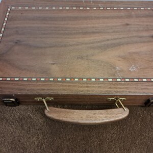 May include: A wooden briefcase with a rectangular design. The case is made of dark brown wood with a decorative border of red, white, and brown rectangles. It has a wooden handle and brass-colored hardware.