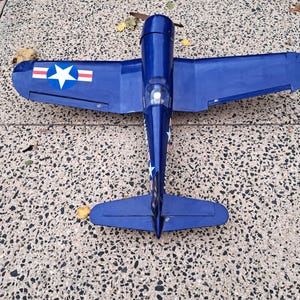 May include: A blue model airplane with white star and stripe decals on the wings and tail. The propeller is brown. The plane is resting on a speckled gray surface.